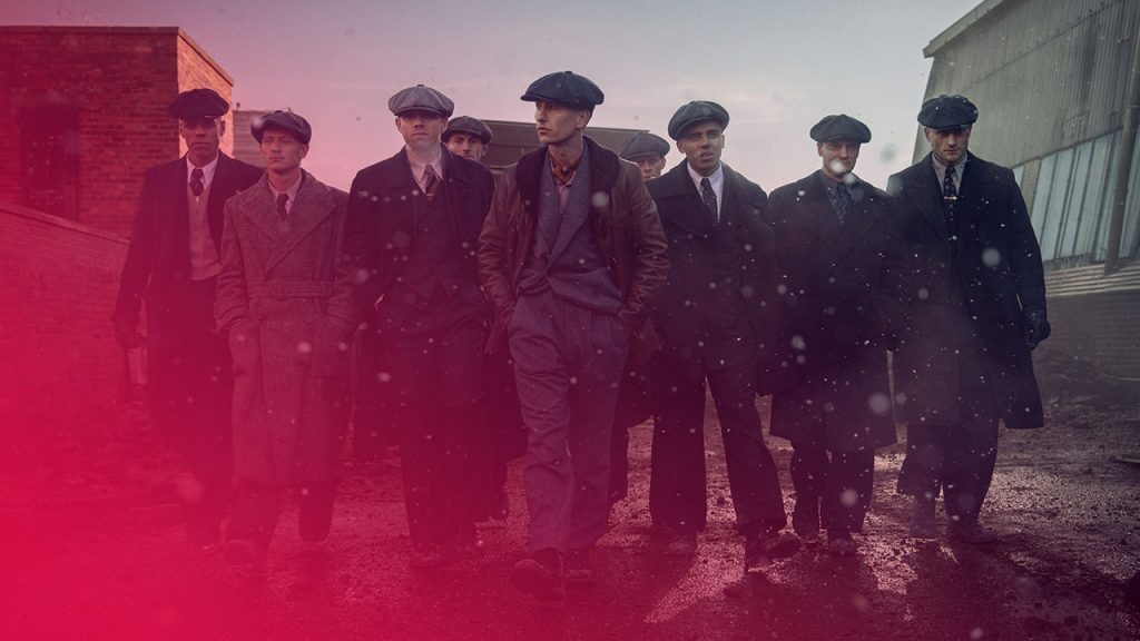 Gruppe von Männern in dunklen Mänteln und Schirmmützen marschiert durch eine industrielle Szenerie bei schneereichem Wetter. Zentrale Figur in brauner Jacke und grauem Anzug führt die Gruppe an.