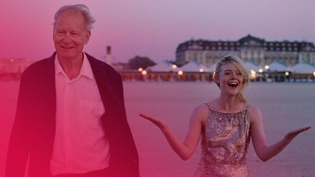 Ein älterer Mann mit grauem Haar und einer jungen Frau mit blonden Haaren stehen am Strand bei Dämmerung. Die Frau trägt ein silbernes Kleid und lacht mit ausgebreiteten Armen. Im Hintergrund sind beleuchtete Strandkörbe und ein großes Gebäude zu sehen. Der Himmel ist in sanften Rosa- und Lilatönen gefärbt.