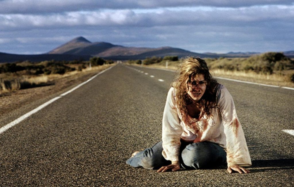Verletzte, verzweifelte Frau kniet auf einsamer Landstraße in karger Berglandschaft; dramatischer, bewölkter Himmel.