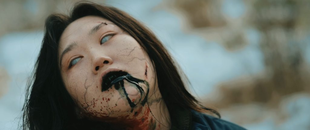 Nahaufnahme einer Frau mit hellen Augen und dunkler Flüssigkeit am Mund, die ihr über das blutverschmierte Kinn läuft.