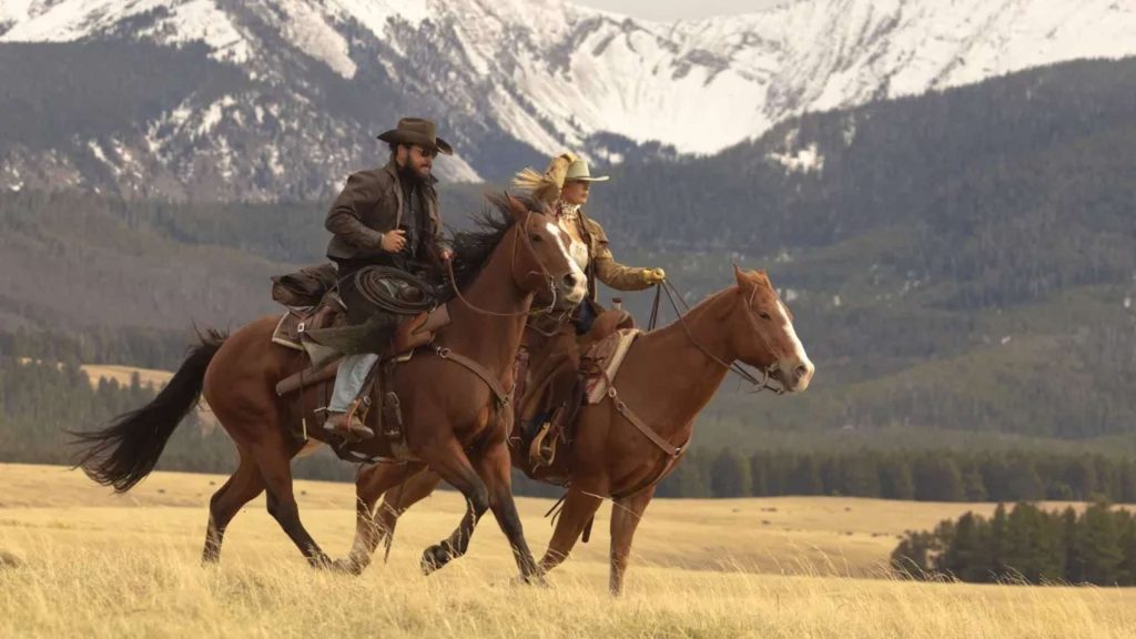 Zwei Reiter, ein Mann mit Hut und Bart und eine Frau mit hellem Hut, galoppieren auf braunen Pferden durch ein goldenes Grasfeld, im Hintergrund schneebedeckte Berge und ein bewaldeter Hügel.