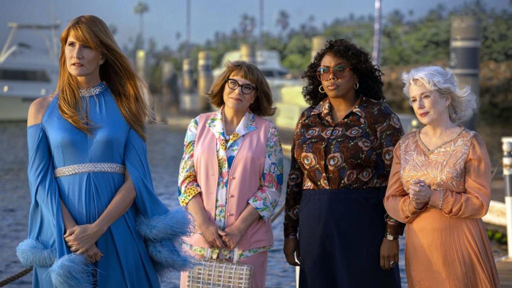 Vier Frauen in farbenfroher 60er-Jahre-Mode stehen nebeneinander an einem sonnigen Hafen vor Yachten.