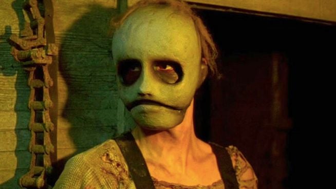 Eine Person mit einer gruseligen, grünlichen Maske und dunklen Augenhöhlen steht in einer düsteren, schattigen Umgebung neben einer rostigen Kette.