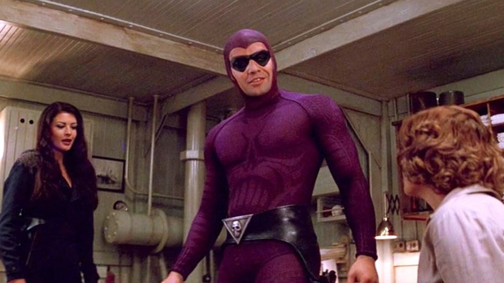 Ein Mann in einem enganliegenden, lila Superheldenkostüm mit Maske und einem Totenkopf-Gürtelschnalle steht zwischen einer Frau und einem Jungen in einem industriell wirkenden Raum.