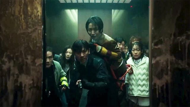 Eine Gruppe von Menschen drängt sich ängstlich in einem düsteren, grünlich beleuchteten Aufzug.