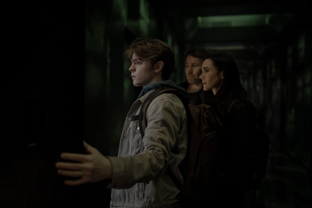 Drei Personen stehen in einem dunklen, grün beleuchteten Raum; der vorderste junge Mann streckt seine Hand aus.