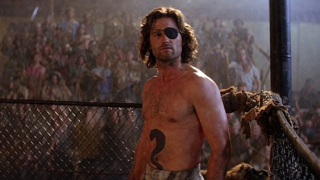 Ein muskulöser Mann mit Augenklappe und einer Tätowierung in Form einer Zwei steht vor einem Zaun, im Hintergrund eine Menschenmenge.