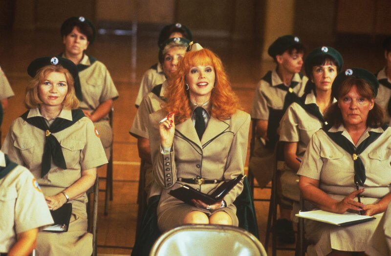 Frau mit roten Haaren in einer beige Uniform sitzt in einem Raum voller Frauen in ähnlichen Uniformen und hält eine Zigarette.