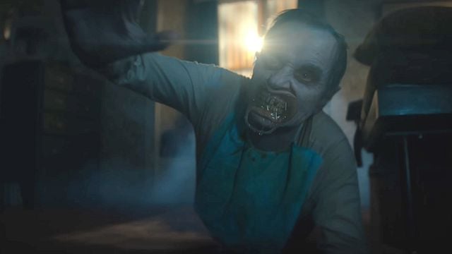 Eine gruselige Figur mit vernarbtem Gesicht und metallischem Mundschutz kriecht in einem düsteren, nebligen Raum auf den Betrachter zu.