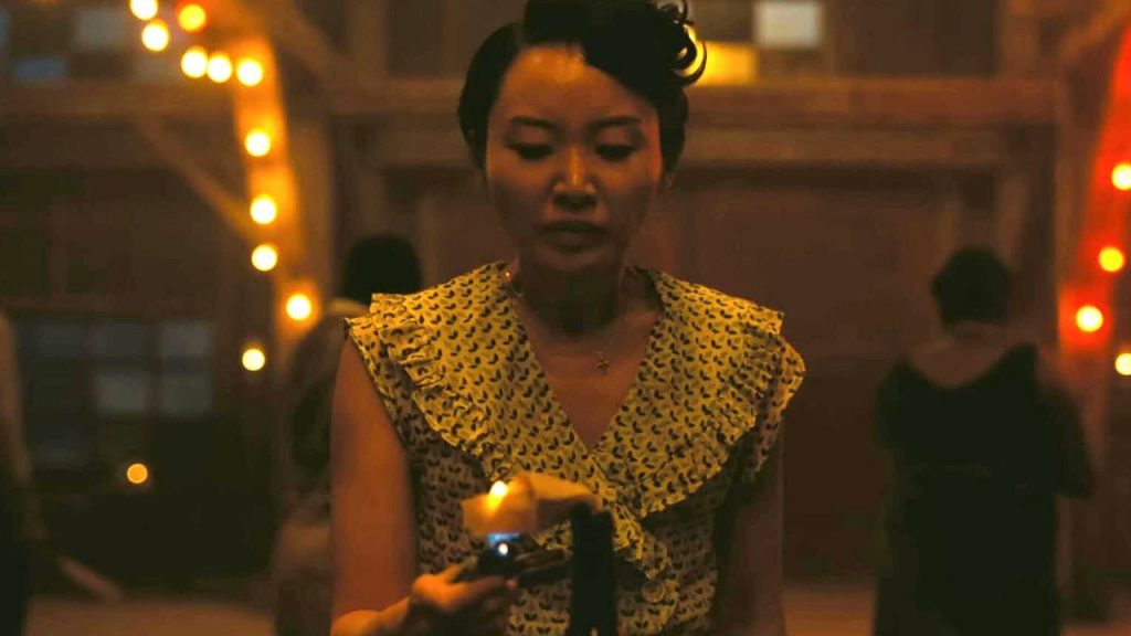 Eine Frau in einem gelben Kleid mit Muster hält ein Feuerzeug in einer warm beleuchteten, festlichen Umgebung bei Nacht.