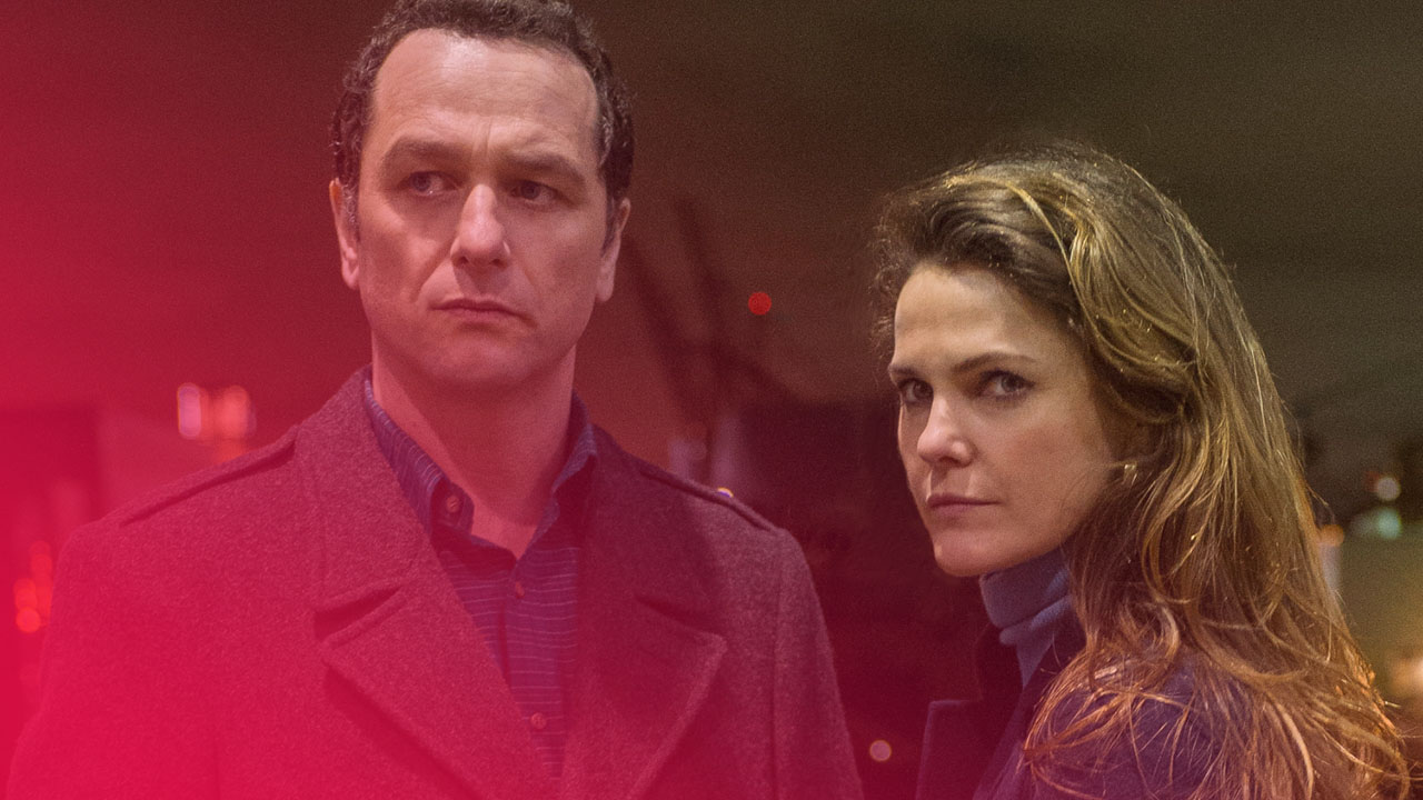 Ein Mann und eine Frau stehen nachts nebeneinander. Beide tragen Mäntel und blicken ernst in die gleiche Richtung. Der Mann hat dunkles Haar und trägt einen grauen Mantel über einem blauen Hemd. Die Frau hat langes, braunes Haar und einen dunkelblauen Mantel. Der Hintergrund ist unscharf und zeigt Lichter einer Stadt bei Nacht.