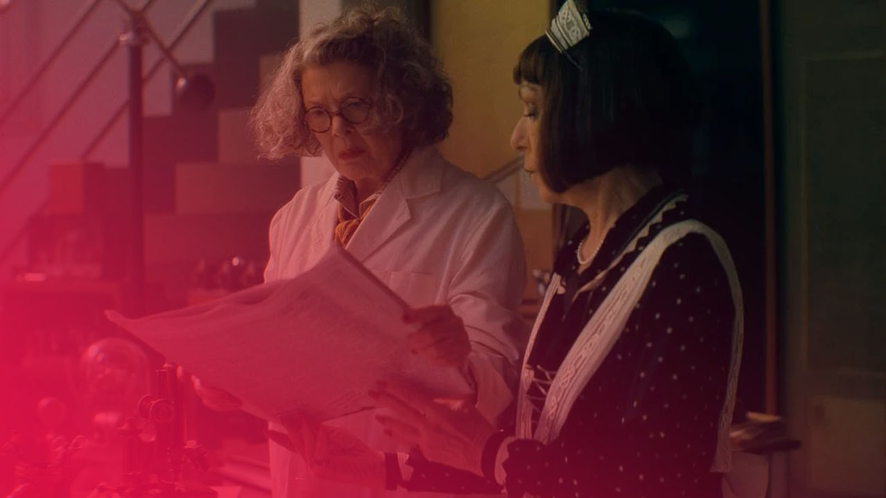 Zwei Frauen stehen in einem Labor oder Arbeitszimmer. Die ältere Frau mit grauen Locken und Brille trägt einen weißen Kittel und hält eine Zeitung. Die jüngere Frau mit dunklem Bob und einer Haarspange trägt ein gepunktetes Kleid mit weißer Schürze. Beide blicken konzentriert auf die Zeitung. Im Hintergrund sind wissenschaftliche Geräte und eine Treppe zu erkennen.