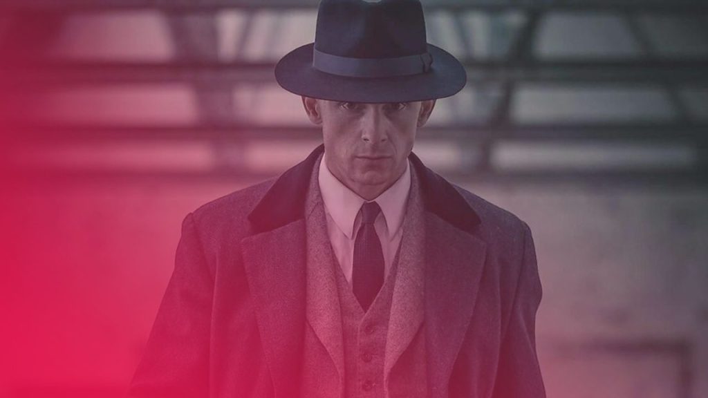 Ein Mann in einem grauen Anzug mit Weste, Krawatte und schwarzem Hut blickt ernst in die Kamera.