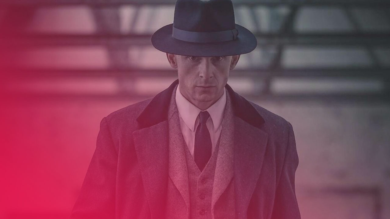 Ein Mann in einem grauen Anzug mit Weste, Krawatte und schwarzem Hut blickt ernst in die Kamera.