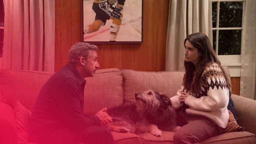 Mann und Frau sitzen auf einem Sofa und schauen sich an; ein struppiger Hund liegt entspannt zwischen ihnen.