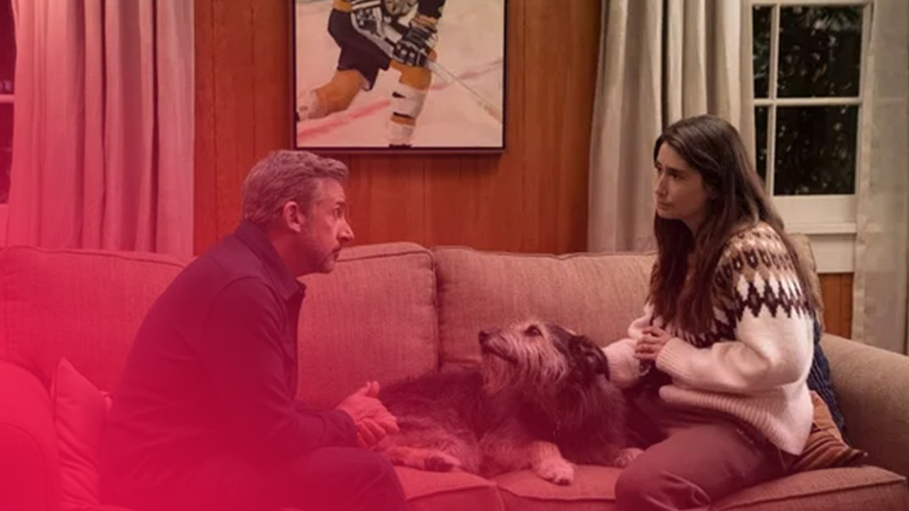 Mann und Frau sitzen auf einem Sofa und schauen sich an; ein struppiger Hund liegt entspannt zwischen ihnen.