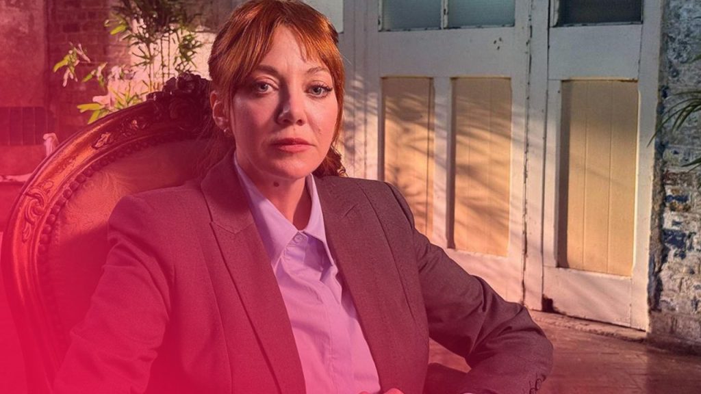 Eine rothaarige Frau in grauem Blazer sitzt auf einem verzierten Sessel, beleuchtet von Sonnenlicht vor einer weißen Tür.