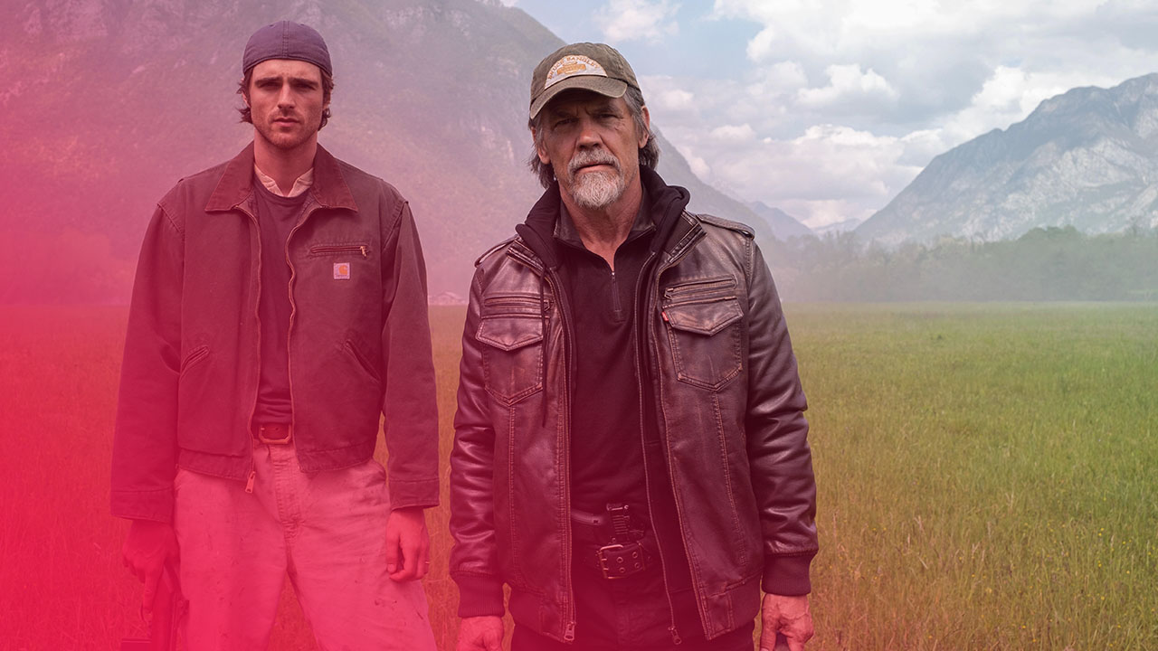 Zwei Männer stehen in einer weiten grünen Wiese vor einer Bergkulisse. Der jüngere Mann links trägt eine grüne Jacke und hält ein Gewehr, während der ältere Mann rechts eine Lederjacke und eine Kappe trägt und einen Kanister hält.