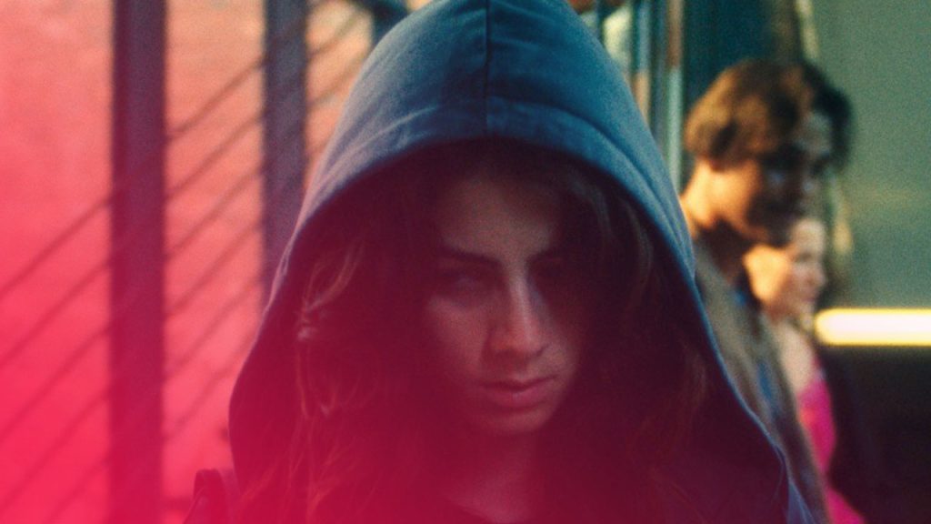 Eine junge Frau mit langen, dunkelbraunen Haaren, die eine dunkle Kapuze über dem Kopf trägt, blickt mit ernstem, fast mürrischem Ausdruck nach unten. Ihr Gesicht ist der zentrale Fokus des Bildes. Im Hintergrund, verschwommen und in einem helleren Bereich, ist eine weitere Person zu erkennen. Die Beleuchtung ist warm und golden, was einen Kontrast zur ernsten Miene der Frau bildet. Das Bild scheint in einer modernen, urbanen Umgebung aufgenommen worden zu sein, möglicherweise in einem Bahnhof oder einer Unterführung, was durch Gitterstrukturen und eine Betonwand im Hintergrund angedeutet wird.