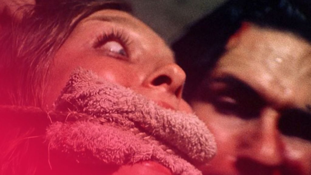 Frau mit Knebel im Mund und weit aufgerissenen Augen; unscharf im Hintergrund ein Mann.