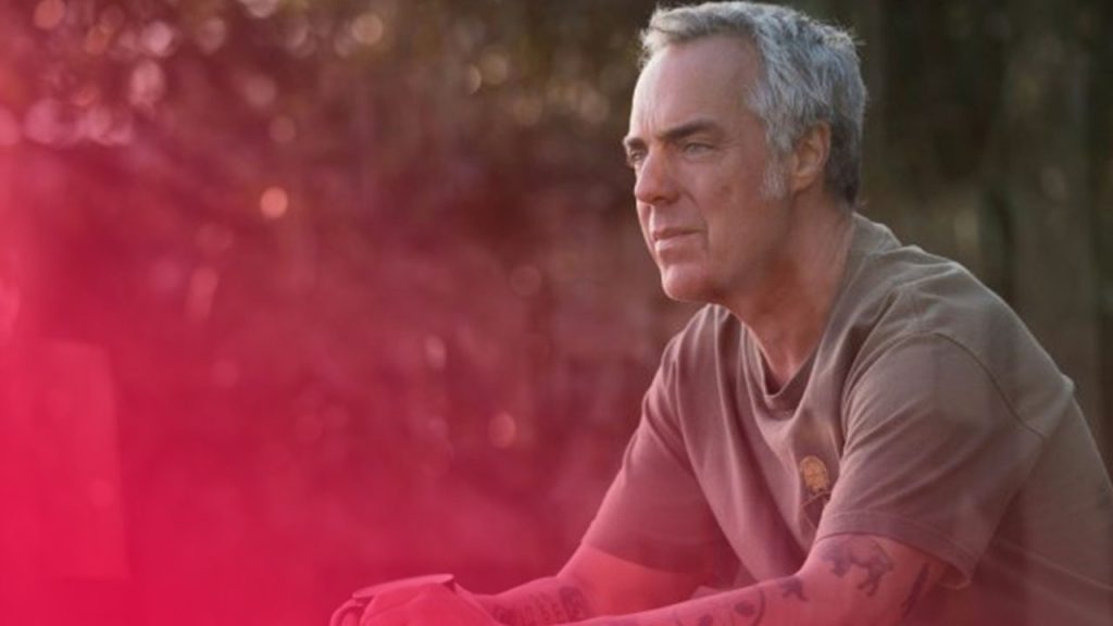 Ein Mann mit grauem Haar und Tattoos an den Unterarmen sitzt im Freien. Er trägt ein olivgrünes T-Shirt und blickt nachdenklich zur Seite. Der Hintergrund ist unscharf und zeigt Bäume im warmen Licht der Sonne.