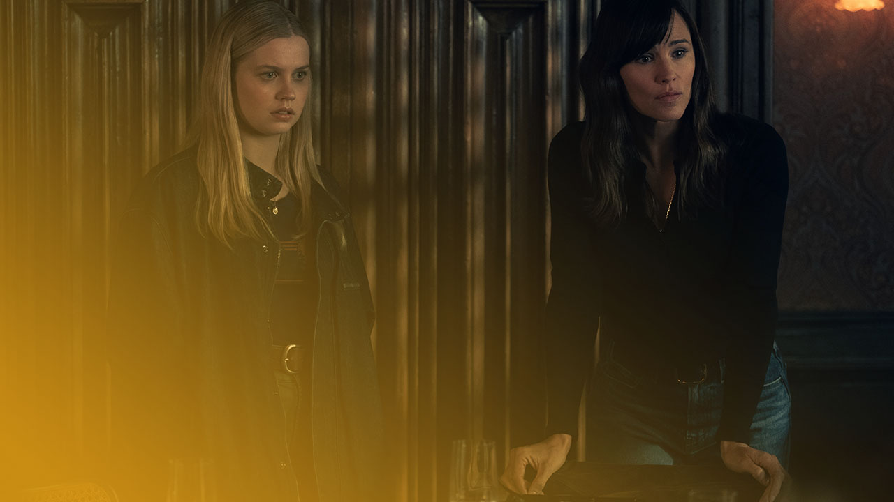 Zwei Frauen stehen in einem düsteren Raum an einem Tisch, beleuchtet von einer einzelnen Lampe, mit besorgten Gesichtsausdrücken.