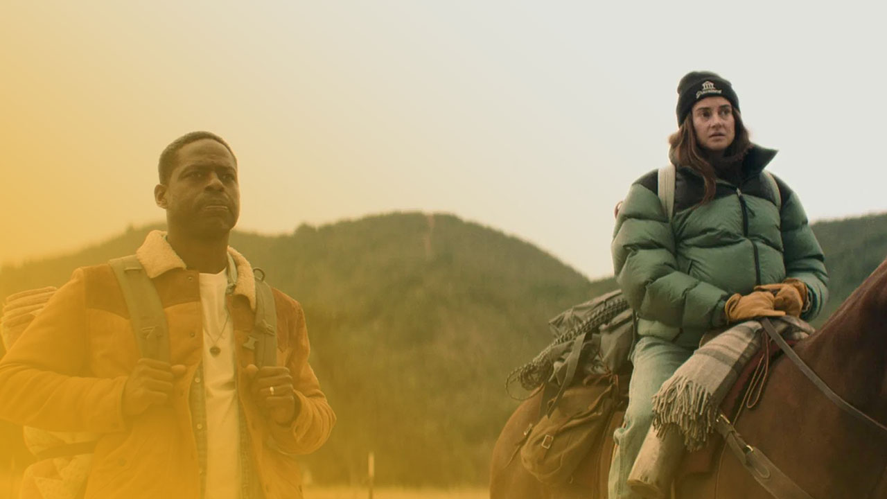 Ein Mann und eine Frau in einer ländlichen Landschaft. Der Mann geht zu Fuß und trägt eine braune Jacke mit Rucksack. Die Frau sitzt auf einem braunen Pferd, trägt eine grüne Daunenjacke und eine schwarze Mütze. Im Hintergrund sind bewaldete Hügel unter einem bedeckten Himmel zu sehen.