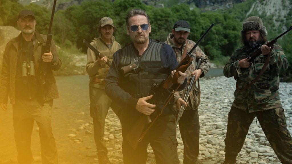 Fünf bewaffnete Männer in Outdoor- und Tarnkleidung stehen bereit in einem flachen Gebirgsfluss, umgeben von grünen Bergen.