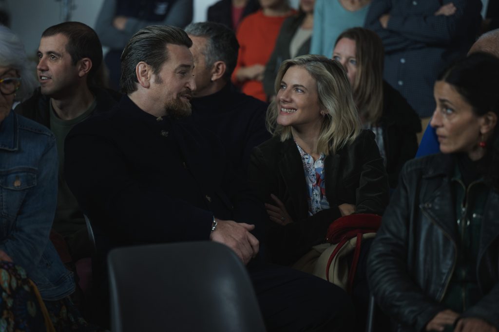 Ein Mann mit Bart und eine lächelnde Frau sitzen nebeneinander in einem Publikum und unterhalten sich während einer Veranstaltung.