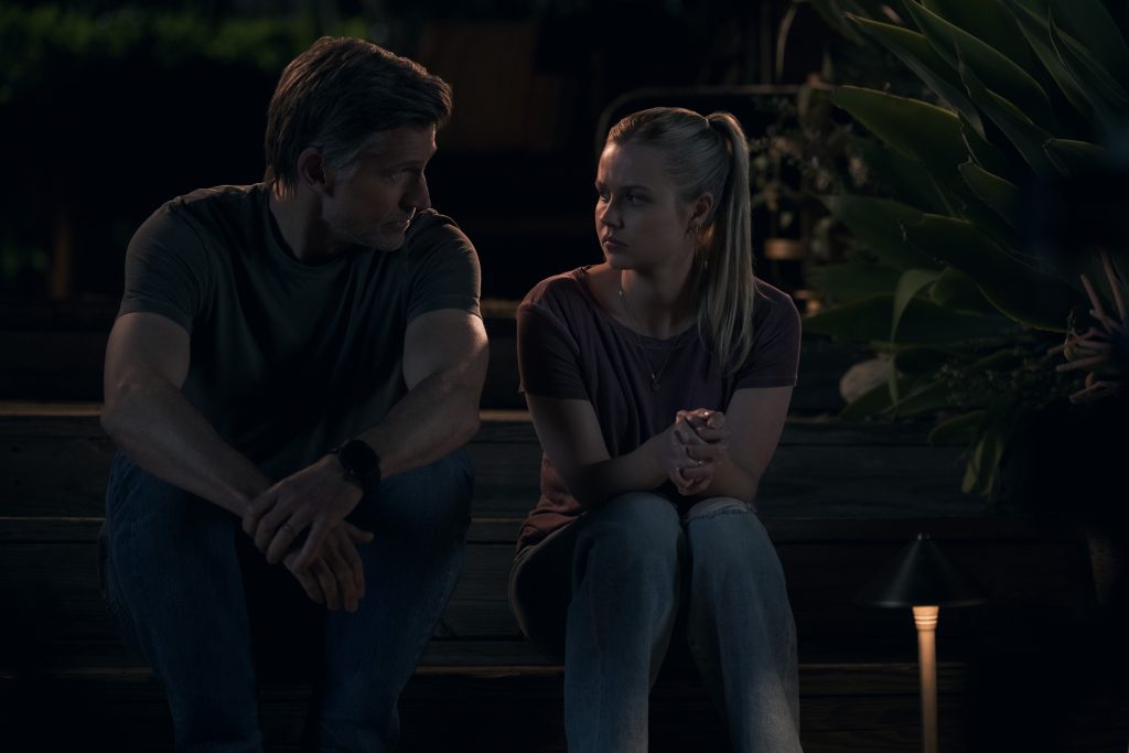 Ein Mann und eine junge Frau sitzen nachts auf einer Holzterrasse und unterhalten sich in gedämpftem Licht.