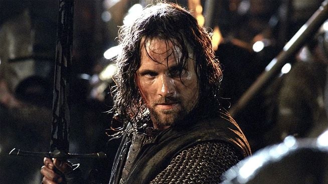 Ein Mann mit langen, dunklen Haaren und Kettenhemd hält in einer düsteren Szene ein Schwert und blickt entschlossen.