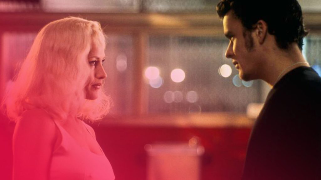 Eine Frau mit blonden Haaren und ein Mann mit dunklem Haar stehen sich in einem schwach beleuchteten Raum gegenüber. Die Frau trägt ein weißes Top, der Mann ein dunkles Oberteil. Sie blicken sich ernst an. Im unscharfen Hintergrund sind Lichter zu sehen, die auf eine Bar oder einen öffentlichen Ort hindeuten.