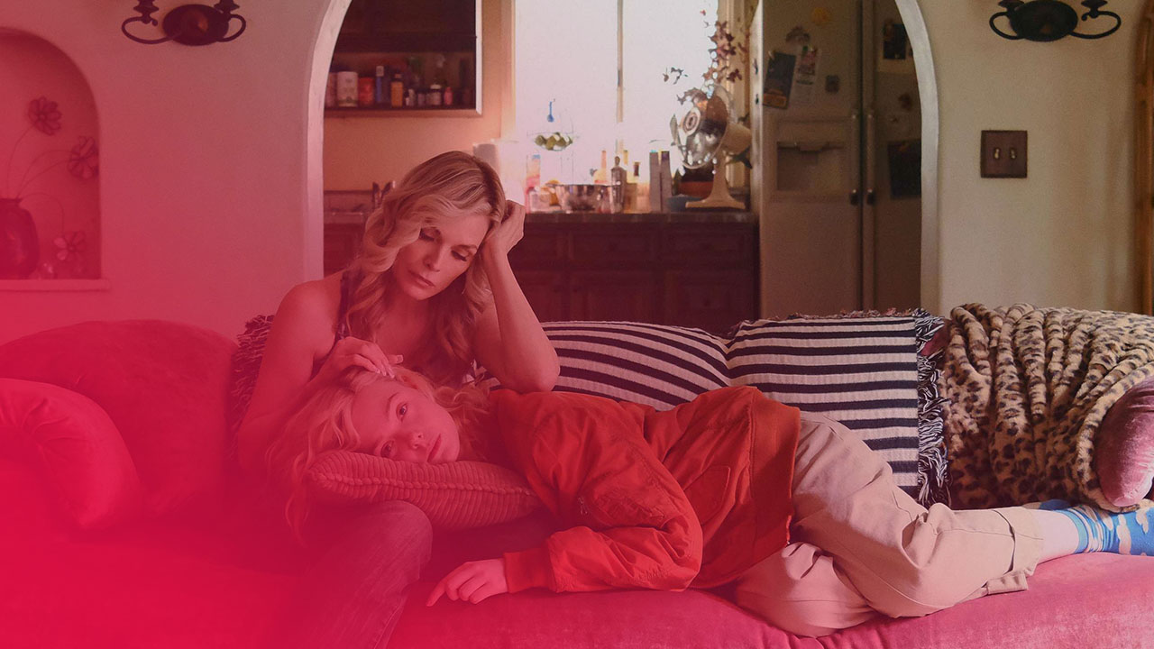 Zwei blonde Frauen auf einer pinken Couch. Die Ältere streichelt sanft den Kopf der Jüngeren, die auf ihrem Schoß ruht. Häusliche Szene.