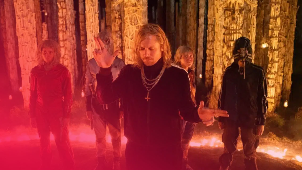 Eine Gruppe von fünf Personen steht vor einem Hintergrund aus hohen, strohbedeckten Pfählen und Feuern. Eine Person in der Mitte trägt einen Trainingsanzug und Ketten mit einem Kreuz, während sie Gesten macht.