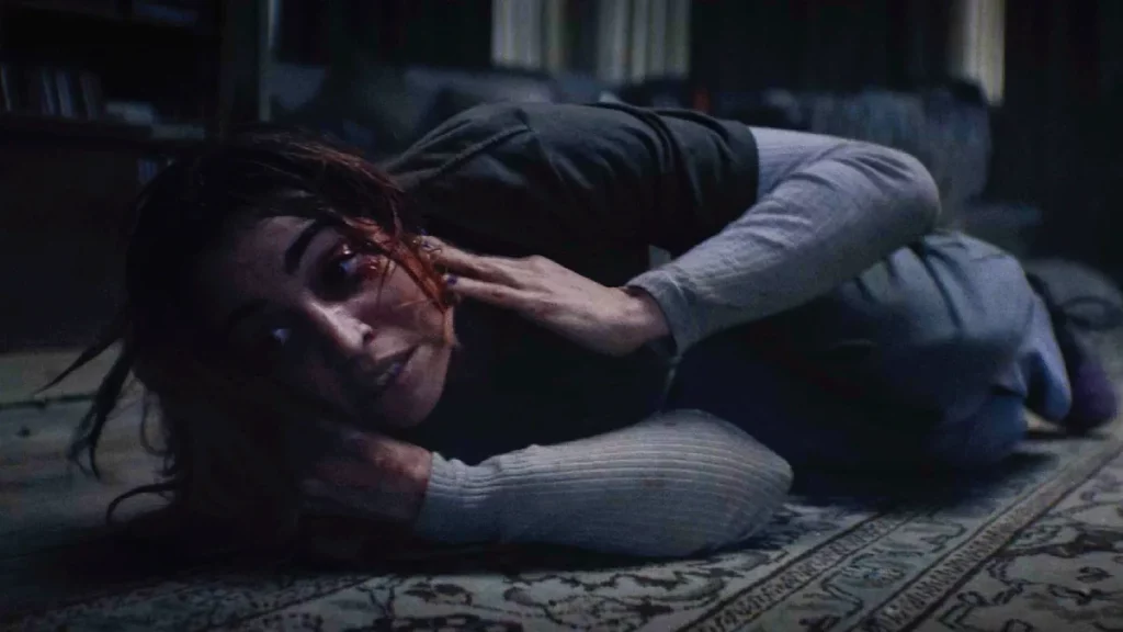 Eine Frau liegt auf dem Bauch auf einem Teppichboden in einem schwach beleuchteten Raum. Ihr Gesicht und ihre Hand sind blutverschmiert, und sie blickt mit schmerzverzerrtem Ausdruck direkt in die Kamera.