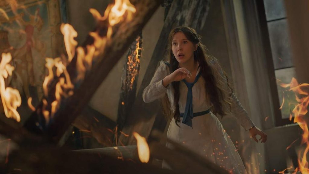 Eine junge Frau in einem weißen Kleid mit blauer Schleife steht erschrocken in einem brennenden Raum, umgeben von Flammen und Funken.