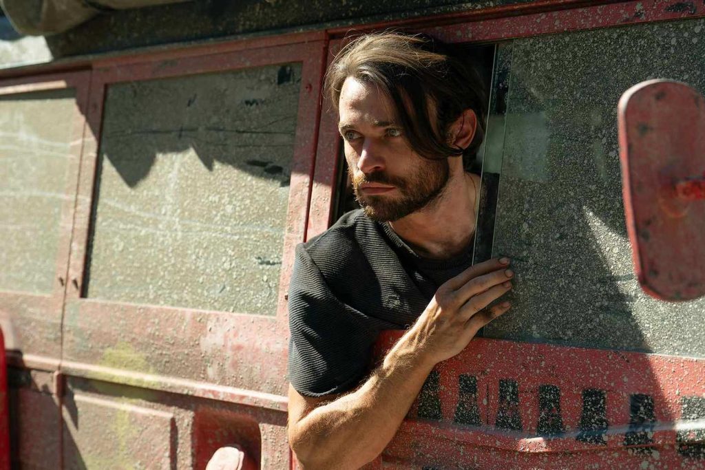 Ein Mann mit Bart und mittellangem Haar schaut aus dem Fenster eines staubigen, roten Fahrzeugs. Er trägt ein dunkles T-Shirt und stützt seinen Arm am Fensterrahmen ab. Die Sonne beleuchtet sein Gesicht und den staubigen Lack des Fahrzeugs.