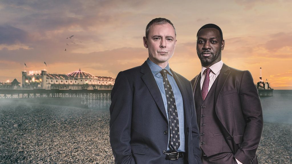 Zwei Männer in Anzügen stehen ernst vor einem beleuchteten Pier am Meer während des Sonnenuntergangs.