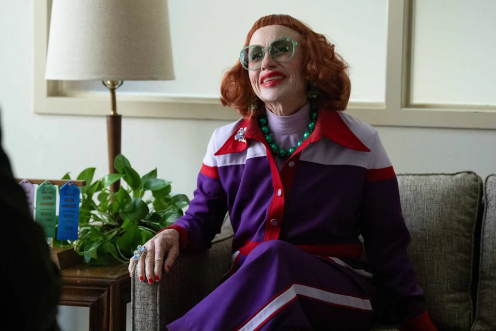 Eine ältere Dame mit roten Haaren und grüner Sonnenbrille sitzt in einem lila-weißen Outfit auf einem Sofa.