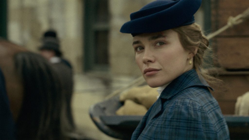 Eine Frau mit einem blauen Hut und einem blau karierten Mantel blickt über ihre Schulter, während im unscharfen Hintergrund ein Pferd und eine historische Szenerie zu erkennen sind.