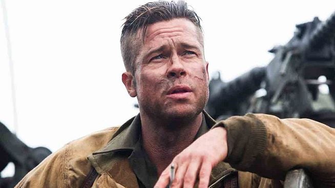 Ein Mann mit schmutzigem Gesicht und kurzer Frisur, bekleidet mit einer braunen Jacke, blickt mit ernstem Ausdruck nach oben, während im Hintergrund unscharf militärisches Gerät zu erkennen ist.