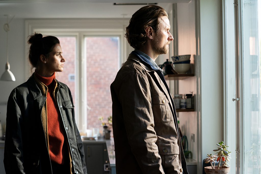 Eine Frau und ein Mann stehen in einer Küche und blicken nachdenklich aus dem Fenster, wobei warmes Tageslicht die Szene beleuchtet.