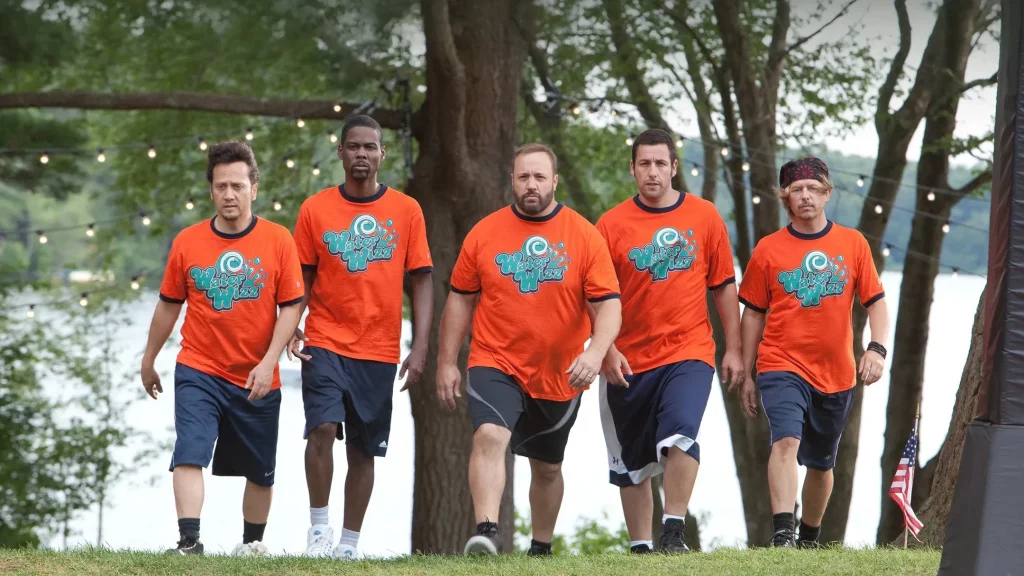 Fünf Männer in identischen orangefarbenen T-Shirts mit dem Aufdruck „Water Wizz“ und dunklen Shorts gehen nebeneinander über eine Wiese, im Hintergrund sind Bäume, ein See und Lichterketten zu sehen.