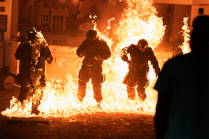 Drei Personen in dunkler Schutzkleidung und Helmen stehen inmitten eines hell lodernden Feuers, das den Boden um sie herum bedeckt, während im Vordergrund die Silhouette einer weiteren Person zu sehen ist.