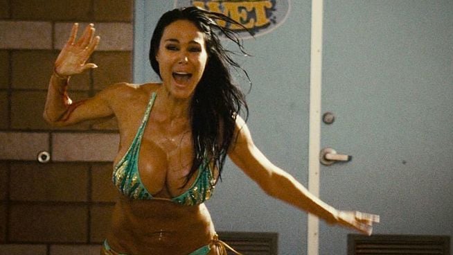 Eine Frau mit langen, dunklen Haaren in einem türkisfarbenen Bikini steht mit erhobenen Armen und offenem Mund da, ihre Haut und Haare sind nass.