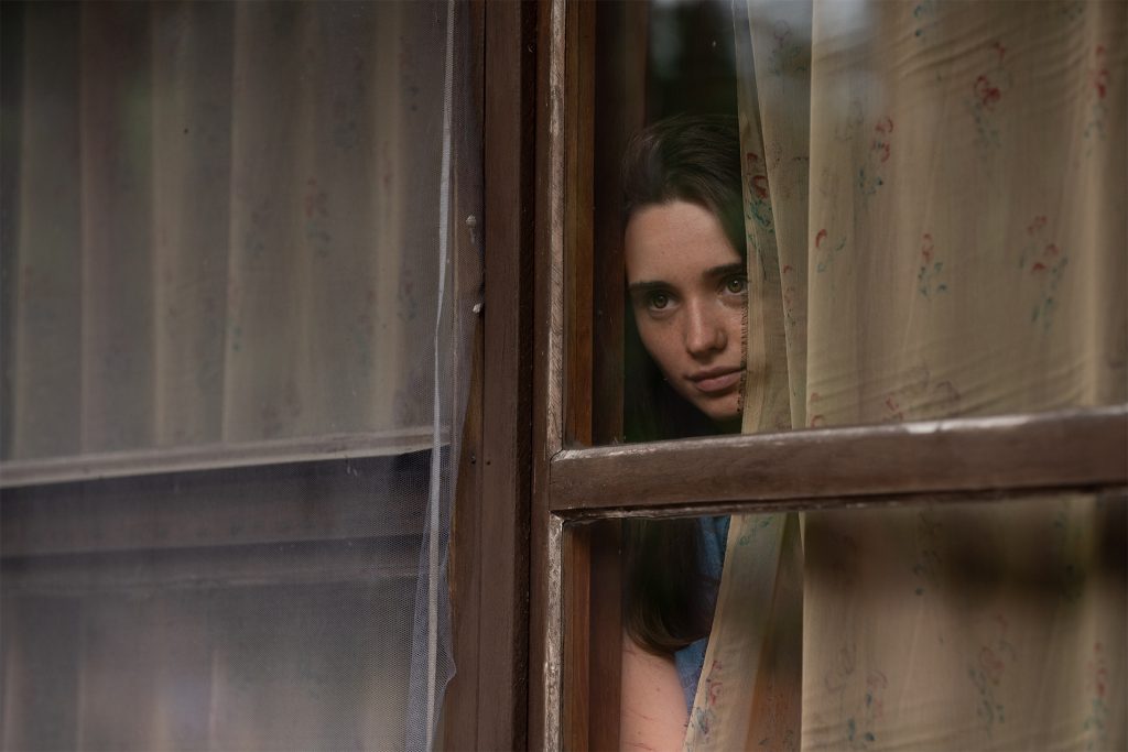Eine junge Frau mit dunklen Haaren blickt ernst und nachdenklich durch ein hölzernes Fenster, wobei ihr Gesicht teilweise von einem dünnen, gemusterten Vorhang verdeckt wird.