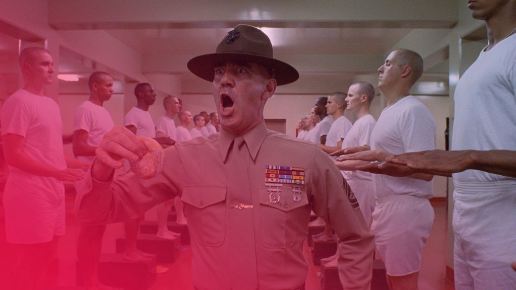 Ein Drill-Instructor in Uniform und mit Hut schreit mit weit aufgerissenem Mund und hält einen Donut in der Hand, während er von Reihen junger Rekruten in weißer Unterwäsche umgeben ist, die in einer Baracke stehen.
