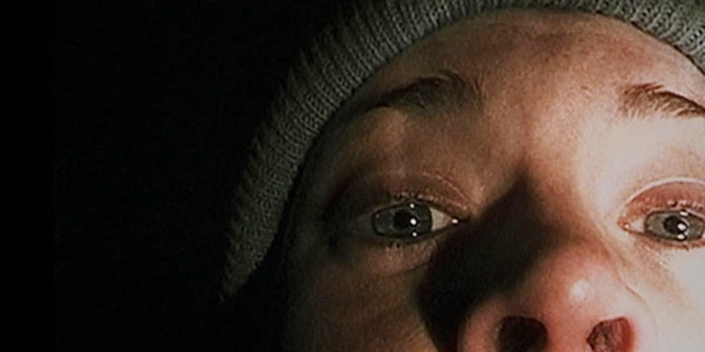 Nahaufnahme eines Gesichts mit weit aufgerissenen Augen und einer grauen Strickmütze, beleuchtet von einer Lichtquelle von unten in dunkler Umgebung.