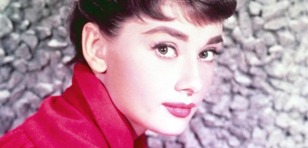 Nahaufnahme von Audrey Hepburn, die mit einem roten Oberteil und dunklem Haar vor einer grauen Steinwand in die Kamera blickt.