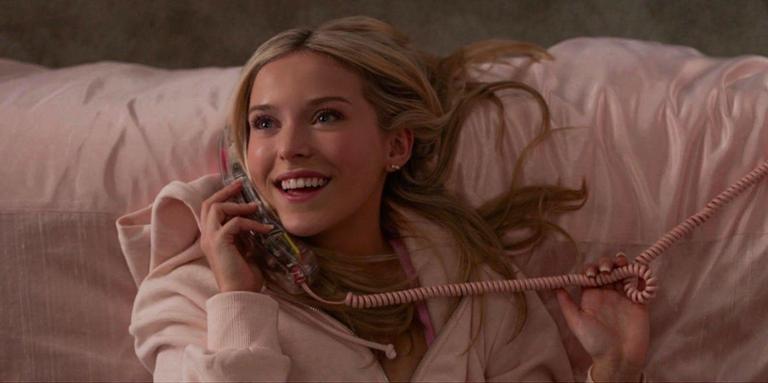Eine junge Frau mit blonden Haaren liegt lächelnd auf einem rosa Bett und telefoniert mit einem rosa Schnurtelefon, das sie in der Hand hält.